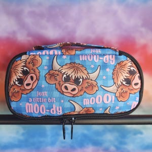 May include: A rectangular zipped pouch featuring a blue plaid pattern with cartoon highland cow heads, small pink stars, and the repeating text "just a little bit moo-dy" and "moo00!" in pink lettering. The case has a black zip and handle.