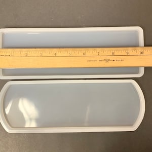 May include: Two clear silicone molds for resin casting. The larger mold is 10 inches long and the smaller mold is 7 inches long. Both molds have a rectangular shape with rounded edges.