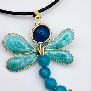 以下が含まれることがあります： 黒いコードが付いたトンボのペンダントネックレス。 トンボは、金色の輪郭を持つターコイズ色の翼、濃い青色のビーズの頭、ビーズの尾が特徴です。 ペンダントは金属とビーズでできています。