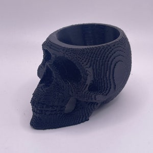 May include: A black, 3D-printed skull-shaped planter. The planter has a hollow interior and is designed with a layered, blocky texture. The skull is angled to the left, showcasing the eye sockets, nose, and jawline.