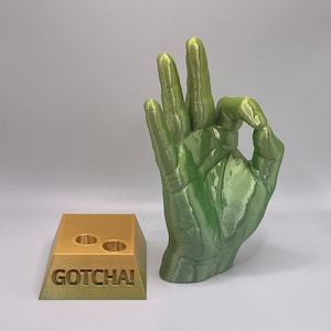 May include: A green 3D printed hand sculpture making the OK sign. The hand is on a gold-colored base with the word "GOTCHA!" printed on it.