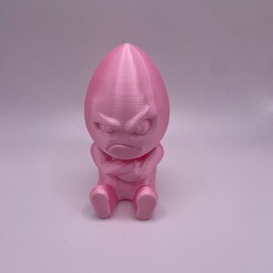 May include: A pink 3D-printed figurine of an egg-shaped character with an angry expression. The figure has arms crossed and is sitting down. The figurine is a solid pink color.