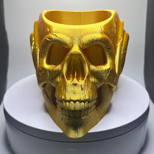 May include: A gold-colored skull planter. The skull is detailed with eye sockets, a nose, and teeth. The planter is made of a smooth, shiny material.