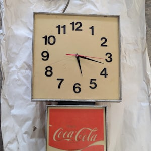 May include: A vintage Coca-Cola clock with a square face and a red and white Coca-Cola sign below. The clock face is cream-colored with black numbers and hands. The clock is powered by a gray cord.