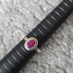 May include: A silver ring featuring an oval, deep red gemstone. The ring is displayed on a black, cylindrical ring sizer against a soft, grey background. The gemstone is the central element, highlighting its rich colour and shape.