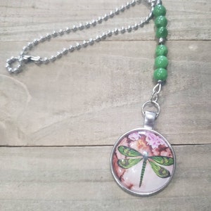 May include: A silver chain with green beads and a round pendant featuring a dragonfly with pink flowers on a brown background.