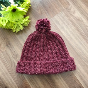 以下が含まれることがあります： 上部にふわふわのポンポンが付いた、ワインレッドのクロシェ編みビーニー