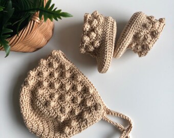 May include: A set of crocheted baby booties and a bonnet in a light beige color. The booties and bonnet have a textured, bumpy design.
