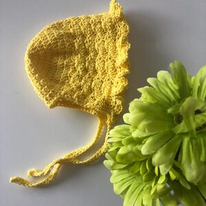 Op de afbeelding: Een gele gehaakte baby muts met een gerimpelde rand en strikjes. De muts ligt op een wit oppervlak naast een groene bloem.