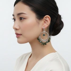 May include: A silver hoop earring featuring a central dark green bead suspended within the circle, with a series of smaller, irregular dark green beads dangling from the bottom edge of the hoop, secured by a silver post and backing mechanism.