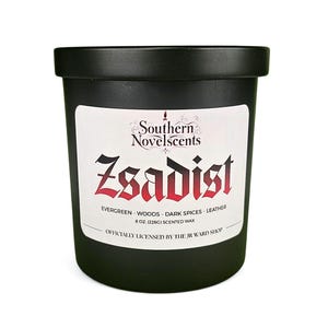 以下が含まれることがあります： 白ラベルの黒いアロマキャンドル。ラベルには「Southern Novelscents」と赤字で「Zsadist」と書かれています。香りのノートとサイズ（226g）も記載されています。