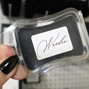 May include: A clear plastic clamshell package containing a black product with gold flecks. A white label with the word "Warden" in a cursive font is affixed to the product. Other similar packages are visible in the background.