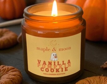 May include: A lit candle in a brown glass jar with a cream-colored label. The label features a crescent moon, a small ghost illustration, and the text maple & moon, VANILLA PUMPKIN COOKIE, and HALLOWEEN EDITION in orange and brown lettering.