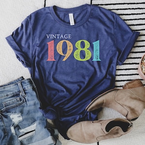 Puede incluir: Una camiseta azul marino con el texto "Vintage 1981" en colores arco&iacute;ris. La camiseta est&aacute; colocada sobre una alfombra de rayas blancas y negras con un par de botines marrones y unos pantalones cortos de mezclilla azul.
