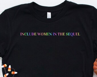 May include: Black t-shirt with the text "INCLUDE WOMEN IN THE SEQUEL" in rainbow colours. The shirt is on a white brick background with a small orange pumpkin and candy corn.