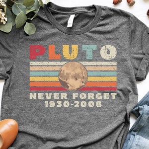 May include: Dark grey heather t-shirt with a vintage-style graphic of Pluto, the text "Pluto Never Forget 1930-2006", and a rainbow stripe design.