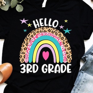 May include: Black t-shirt with a rainbow and leopard print design. The rainbow has a pink heart in the center. The text "Hello 3rd Grade" is printed below the rainbow. The shirt is on a white surface.