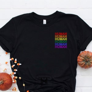 Puede incluir: Camiseta negra con un diseño de arcoíris que dice "HUMAN" repetido cinco veces.