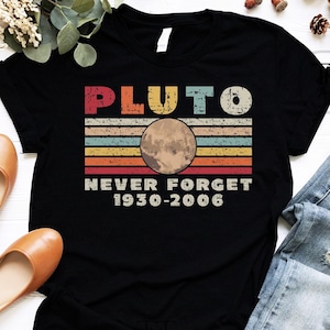 May include: Black t-shirt with a retro design featuring the word "PLUTO" above a graphic of the planet and the text "NEVER FORGET 1930-2006". The design has a vintage, distressed look.