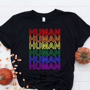 Puede incluir: Camiseta negra con la palabra "HUMAN" repetida seis veces en colores arcoíris. La palabra "HUMAN" está delineada en colores neón.