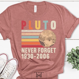 May include: A heather mauve t-shirt with a retro-style graphic that says "Pluto Never Forget 1930-2006". The graphic features a stylized image of Pluto with colorful stripes behind it.