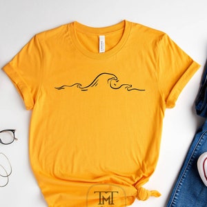 Könnte beinhalten: Ein gelbes T-Shirt mit kurzen Ärmeln und einer schwarzen Linienzeichnung einer Welle auf der Vorderseite. Das Shirt ist unten verknotet. Das Wort "Gold" ist in Schwarz unter dem Shirt gedruckt.