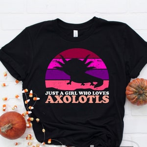 Könnte beinhalten: Schwarzes T-Shirt mit einem pinkfarbenen und violetten Sonnenuntergangs-Motiv, das eine schwarze Axolotl-Silhouette zeigt. Der Text "Just a girl who loves axolotls" ist in Pink unter dem Motiv gedruckt.
