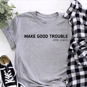 Pode incluir: Camiseta cinza com o texto "MAKE GOOD TROUBLE" e "JOHN LEWIS" impresso nela. A camiseta está colocada sobre um fundo de tijolo branco com uma camisa xadrez branca e preta e tênis pretos.