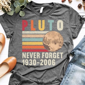 May include: A dark grey heather t-shirt with a vintage-style graphic of Pluto, a planet with rings, and the text "Pluto Never Forget 1930-2006".