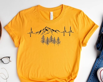 Könnte beinhalten: Ein gelbes T-Shirt mit einem schwarzen Berg- und Kiefernwald-Motiv. Das Motiv enthält eine Herzschlaglinie. Der Text "Gold" ist unter dem Motiv gedruckt.