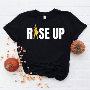 May include: Black t-shirt with a white graphic of a person raising their arms in the air with the text "RISE UP" in white.