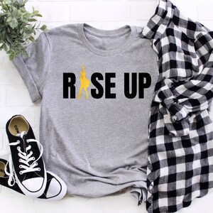 May include: Gray t-shirt with the text "RISE UP" in black letters and a yellow silhouette of a person raising their arms in the air.