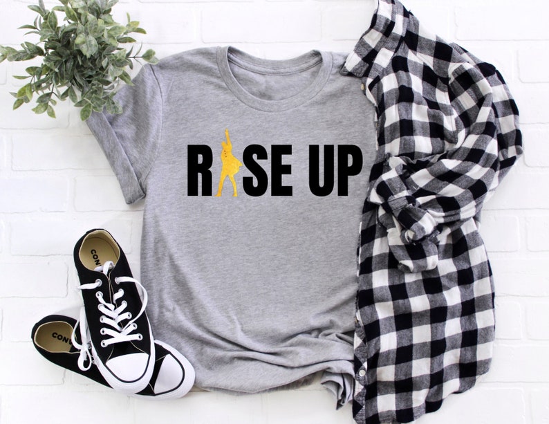 May include: Gray t-shirt with the text "RISE UP" in black letters and a yellow silhouette of a person raising their arms in the air.