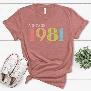 Puede incluir: Una camiseta de color rojo heather con el texto "Vintage 1981" en colores arco&iacute;ris. Los n&uacute;meros est&aacute;n desgastados y tienen un aspecto vintage.
