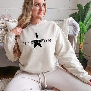 May include: A cream-colored sweatshirt with a black silhouette of a person standing on a star with the word "HAMILTON" printed below.