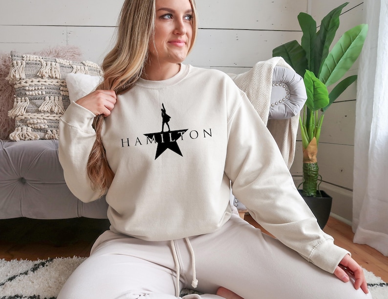 May include: A cream-colored sweatshirt with a black silhouette of a person standing on a star with the word "HAMILTON" printed below.