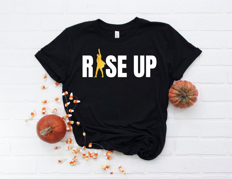 May include: Black t-shirt with a white graphic of a person raising their arms in the air with the text "RISE UP" in white.