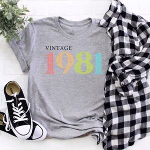 Puede incluir: Camiseta gris con el texto "Vintage 1981" en colores arco&iacute;ris. El a&ntilde;o est&aacute; en una fuente desgastada.