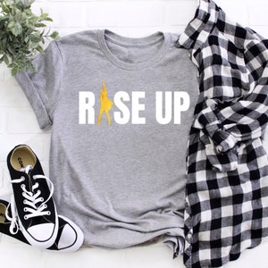 May include: Gray t-shirt with the text "RISE UP" in white letters. A yellow silhouette of a person is inside the "R".
