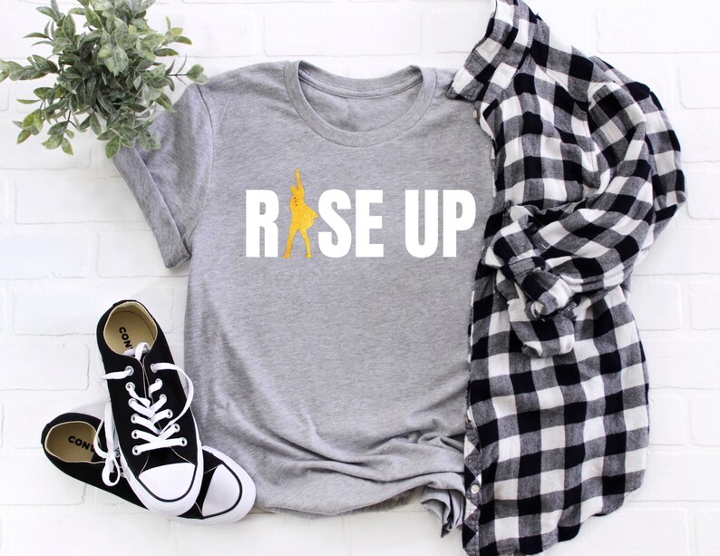 May include: Gray t-shirt with the text "RISE UP" in white letters. A yellow silhouette of a person is inside the "R".