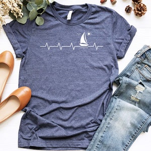 May include: A navy blue t-shirt with a white sailboat graphic and a heartbeat line. The graphic also includes a small sun and waves. The shirt is laid flat on a white surface with a pair of brown flats and blue jeans.