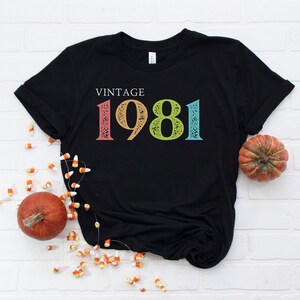 Puede incluir: Camiseta negra con el texto "Vintage 1981" en colores arco&iacute;ris. La camiseta est&aacute; sobre un fondo de ladrillos blancos con calabazas y ma&iacute;z dulce.