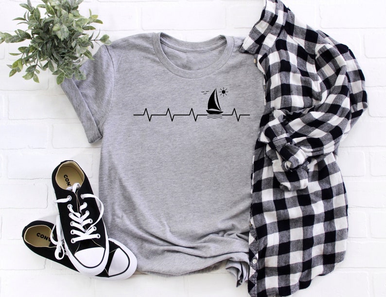May include: A gray t-shirt with a black sailboat and sun graphic. The graphic is a stylized heartbeat with a sailboat and sun on the peak of the heartbeat.
