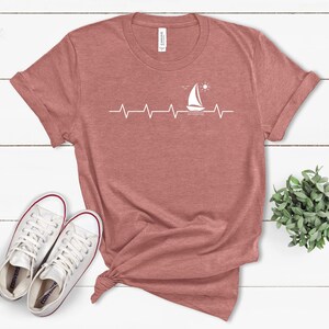 May include: A maroon t-shirt with a white sailboat graphic and a heartbeat line. The sailboat is sailing on a wavy line with a sun above it.