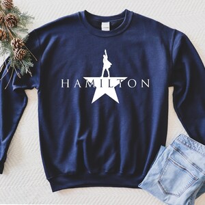 May include: A navy blue sweatshirt with a white silhouette of a person standing on a star with the word "HAMILTON" printed below.