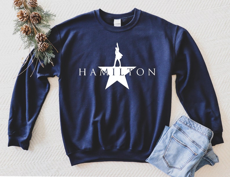 May include: A navy blue sweatshirt with a white silhouette of a person standing on a star with the word "HAMILTON" printed below.