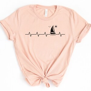 May include: A light pink t-shirt with a black graphic of a sailboat, sun, and birds on a heartbeat line.