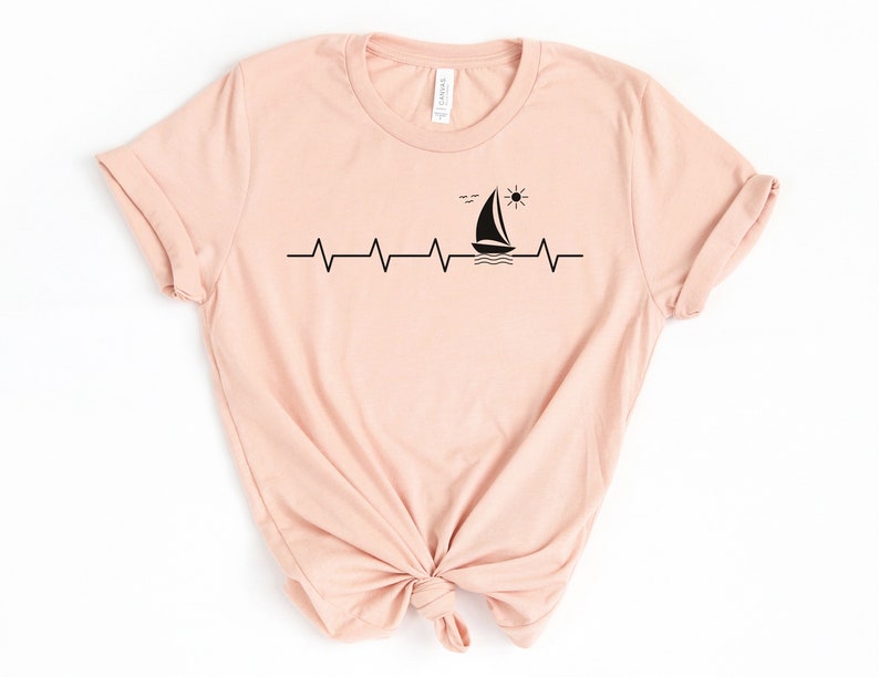 May include: A light pink t-shirt with a black graphic of a sailboat, sun, and birds on a heartbeat line.