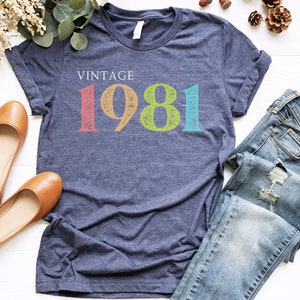 Puede incluir: Una camiseta azul marino con el texto "Vintage 1981" en letras coloridas y desgastadas.