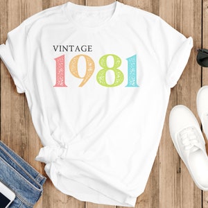 Puede incluir: Camiseta blanca con el texto "Vintage 1981" en colores arco&iacute;ris. Los n&uacute;meros est&aacute;n desgastados y tienen un aspecto vintage.